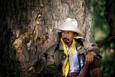 Man wearing hat while sitting against tree trunk in forest