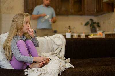 Little girl is sick, coughs, sitting in the living room on the couch, dad pours water to his child