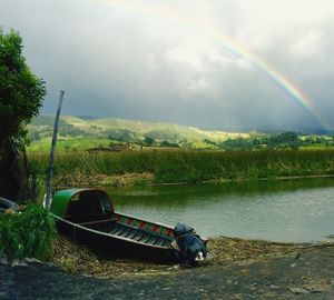 Boat moored at lakeshore against rainbow below cloudy sky