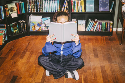Full length of young woman reading book while sitting cross-legged in bookstore