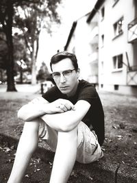 Portrait of mid adult man wearing eyeglasses sitting on footpath against buildings