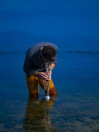 Man playing trumpet while standing in sea against sky at dusk