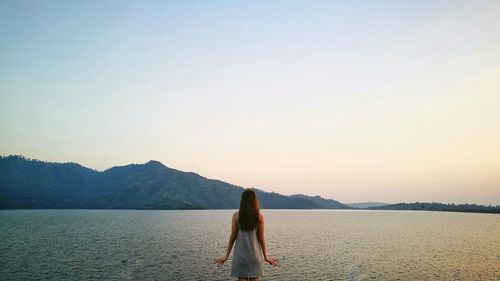 Rear view of woman looking at sea while standing against clear sky during sunset