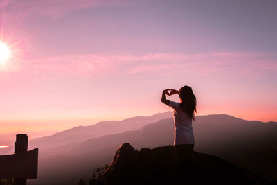Rear view of woman on mountain making heart shape against sky during sunset