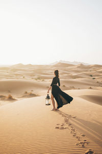 Side view of woman holding lantern while standing at sahara desert against clear sky during sunset