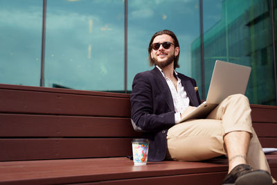 Photo of male freelancer with laptop sitting on wooden bench next to glass wall