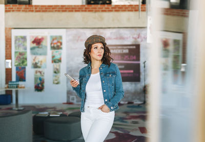 Charming young woman in jeans jacket using mobile phone in modern art gallery exhibition