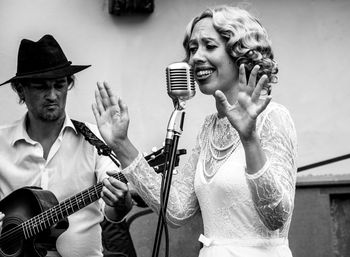 Woman singing on microphone with man playing guitar against wall