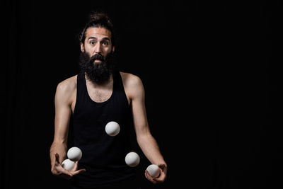 Portrait of bearded man playing with balls while standing against black background