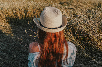 Rear view of woman wearing hat sitting amidst wheat crops at sunset