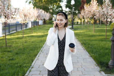 Young woman holding coffee talking over smart phone while standing on footpath
