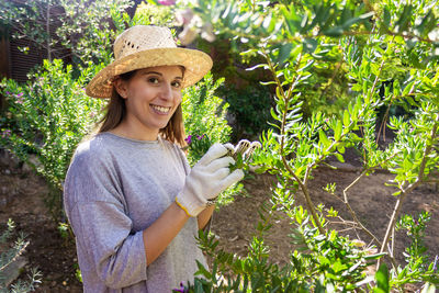 Portrait of smiling young woman wearing hat touching plants in garden