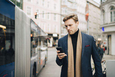 Male entrepreneur using internet through mobile phone while standing by bus in city