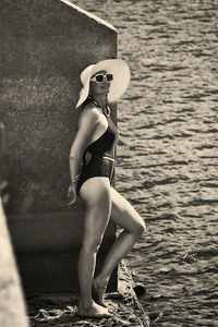 Portrait of a woman in a swimsuit, hat and sunglasses in summer on the riverbank by a concrete wall