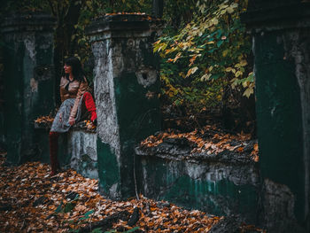 Full length of woman sitting on damaged retaining wall by fallen autumn leaves