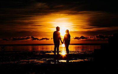 Silhouette men standing on shore against orange sky