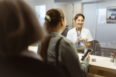 Happy female receptionist looking at woman through glass window at hospital