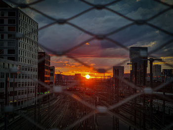 Cityscape seen through chainlink fence against sky during sunset