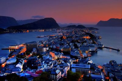 Illuminated cityscape by sea against sky at sunset