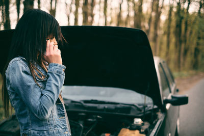 Side view of woman using phone while standing by car in forest