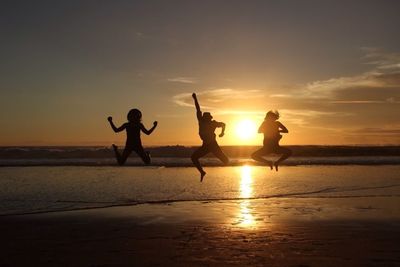 Silhouette friends jumping at beach against sky during sunset