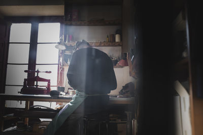 Back view of unrecognizable craftsman sitting working in dark workshop