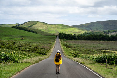 Rear view of woman with yellow coat walking on a long, straight road with mountains in background