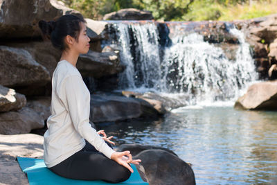 Side view of woman doing lotus position yoga while sitting on rocks against waterfall