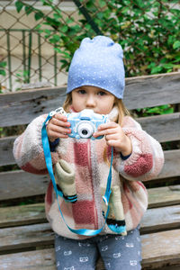 A young girl is sitting on a rustic wooden bench in a park, taking pictures with a blue camera. 