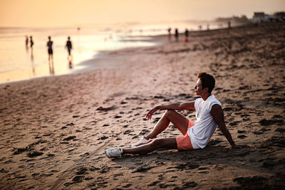 Rear view of man sitting on shore at beach during sunset