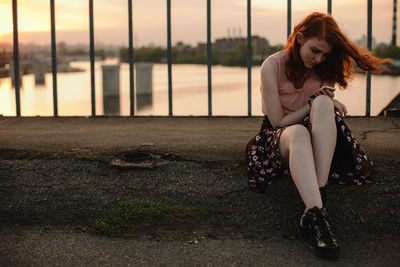 Young redhead woman using smart phone while sitting on bridge in city