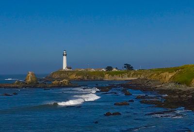 Lighthouse by sea against clear blue sky