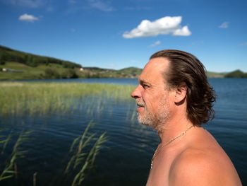 An unshaven adult man stands in profile against the backdrop of the lake shore on a summer sunny day