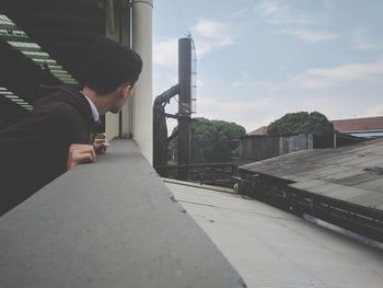 Side view of young man sitting on built structure against sky