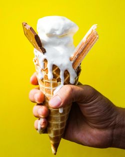 Close-up of hand holding ice cream cone against yellow background