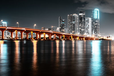 Illuminated bridge over river by buildings against sky at night