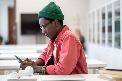 African student guy sitting at table in library holding smartphone taking break from studying