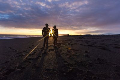 Silhouette couple holding hands while standing at beach against cloudy sky during sunset