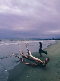 Side view of woman standing by driftwood at beach against cloudy sky