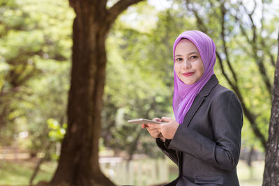 Portrait of young woman wearing hijab using mobile phone while sitting in park