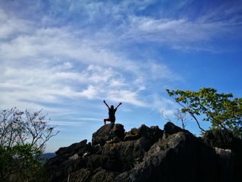 Low angle view of female hiker with arms raised standing on rocky mountain against blue sky