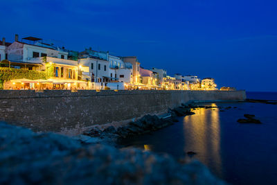 Illuminated buildings by sea against blue sky at dusk