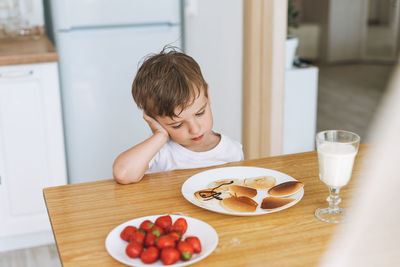 Cute toddler boy sitting with puncakes with berries in kitchen at home. child doesn't want to eat