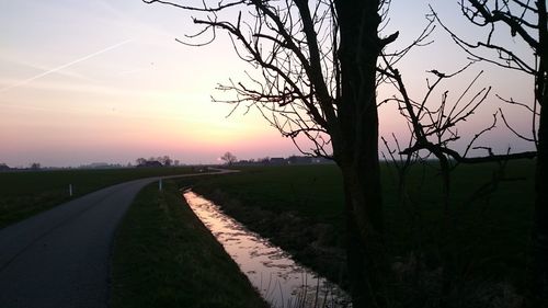 Road amidst bare trees on field against sky at sunset