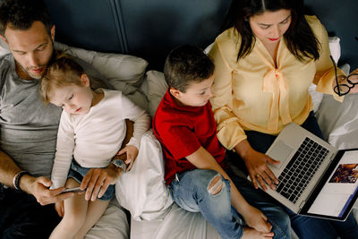 Directly above shot of parents using technologies while sitting with children on bed