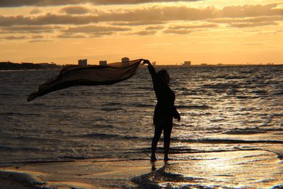 Silhouette woman holding shawl while standing on sea shore against sky during sunset