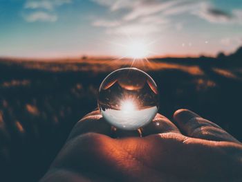 Cropped hand of person holding crystal ball against sky during sunset