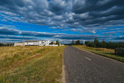 Empty road by grassy field against cloudy sky