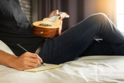 Midsection of man writing on spiral notebook while playing guitar at home