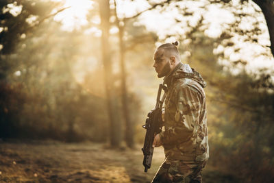 Side view of army soldier holding gun standing in forest